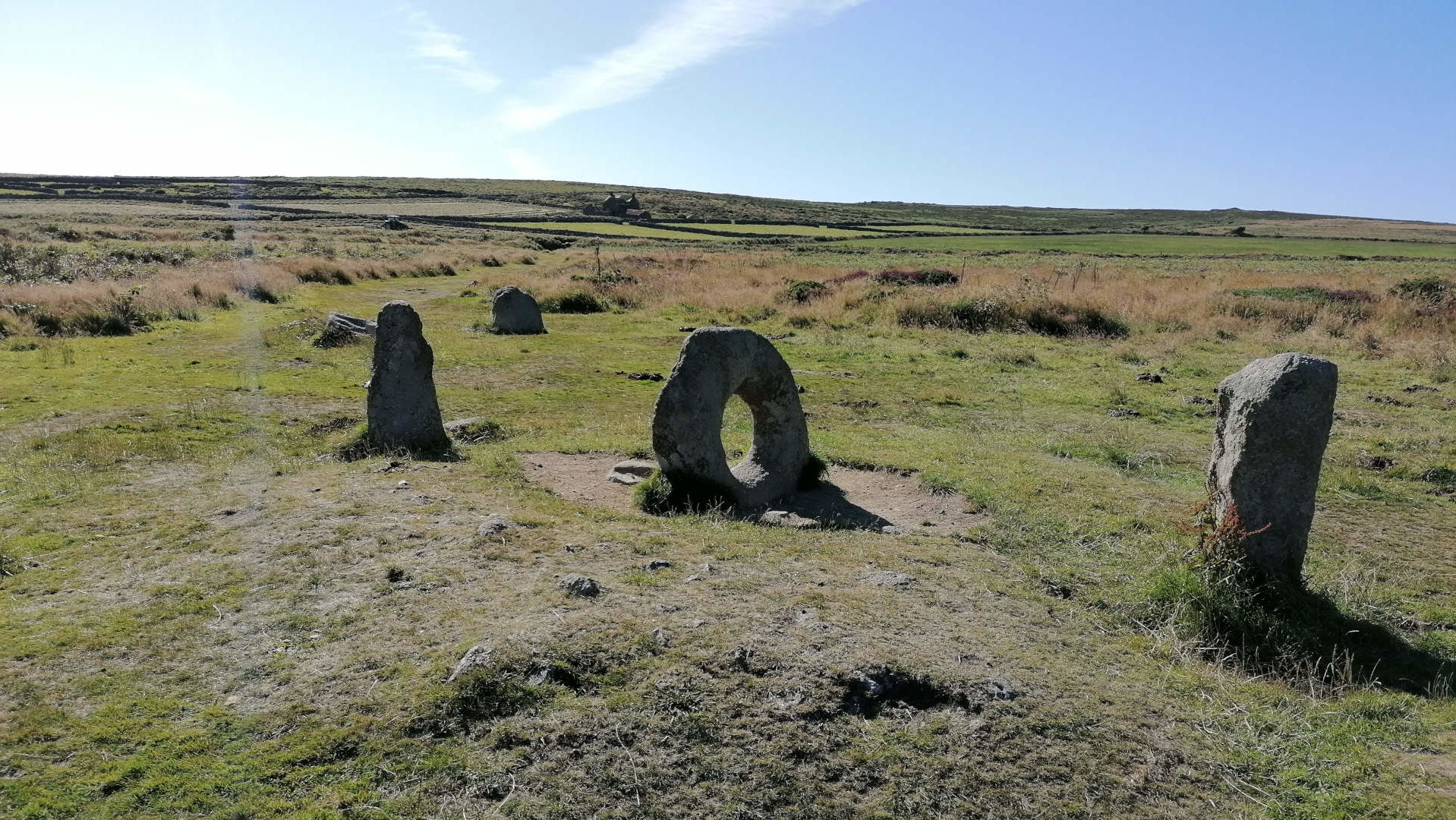 A grassy field under a bright blue sky. Several large, upright stones, some shaped like pillars and one appearing as a ring, are scattered across the field. The stones appear to be part of some sort of ancient stone arrangement, possibly a ruin or a prehistoric monument. The overall impression is one of a remote, historical site in a natural, open landscape.