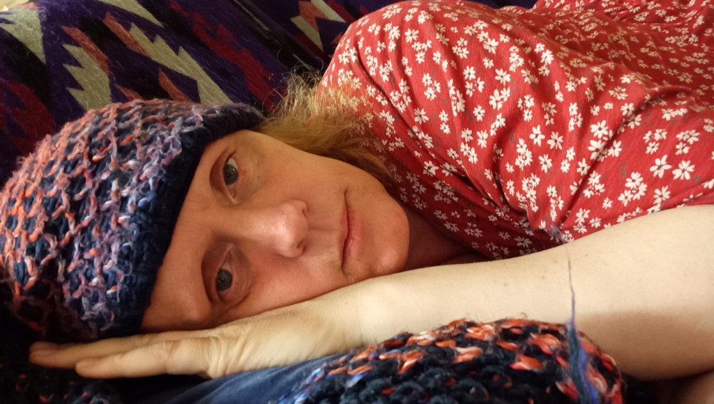 Close-up of a fair-skinned Leonie lying down, possibly resting or sleeping. They are wearing a red floral-print top and a multicoloured knitted hat. Their expression appears somewhat pensive or melancholic. The background features a patterned blanket or throw. The overall mood is quiet and introspective.