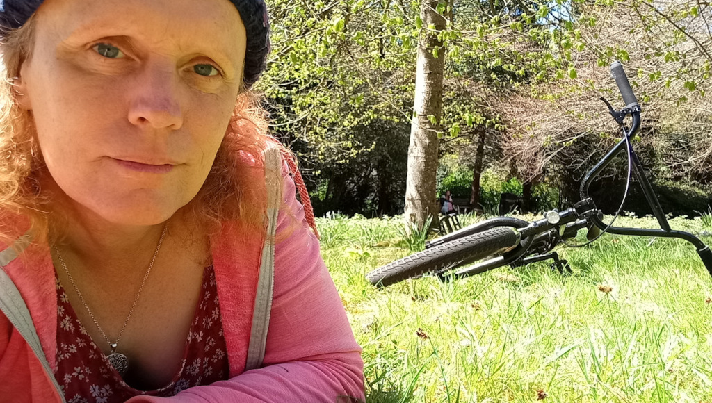Leonie  lying on the grass in a park. She's wearing a pink jacket and a blue hat, and has reddish-blonde hair. She is looking directly at the camera. Behind her, a bicycle is resting on the grass, and there are trees and some indistinct figures in the background suggesting other people are present in the park. The overall impression is one of relaxed contentment in a peaceful outdoor setting.