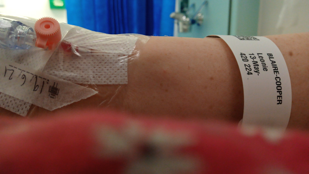 Close-up of Leonie's arm in a hospital setting. A cannula is visible, taped to the arm, along with a hospital identification band. The band indicates the patient's name (Leonie Blaire-Cooper) and hospital information. The background is blurred but shows a typical hospital environment. The overall impression is one of medical treatment or hospitalisation.