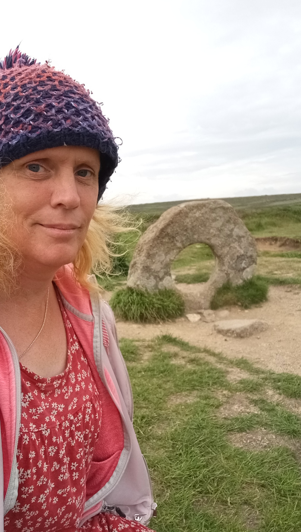 Selfie of Leonie wearing a purple and pink knitted hat and a red floral dress under a light gray zip-up jacket. She is standing in front of a large, ring-shaped stone structure, which appears to be outdoors in a grassy area. The sky is cloudy and the overall mood conveys a sense of peaceful discovery or exploration. Leonie's expression is calm and pleasant, suggesting a positive experience at the site.