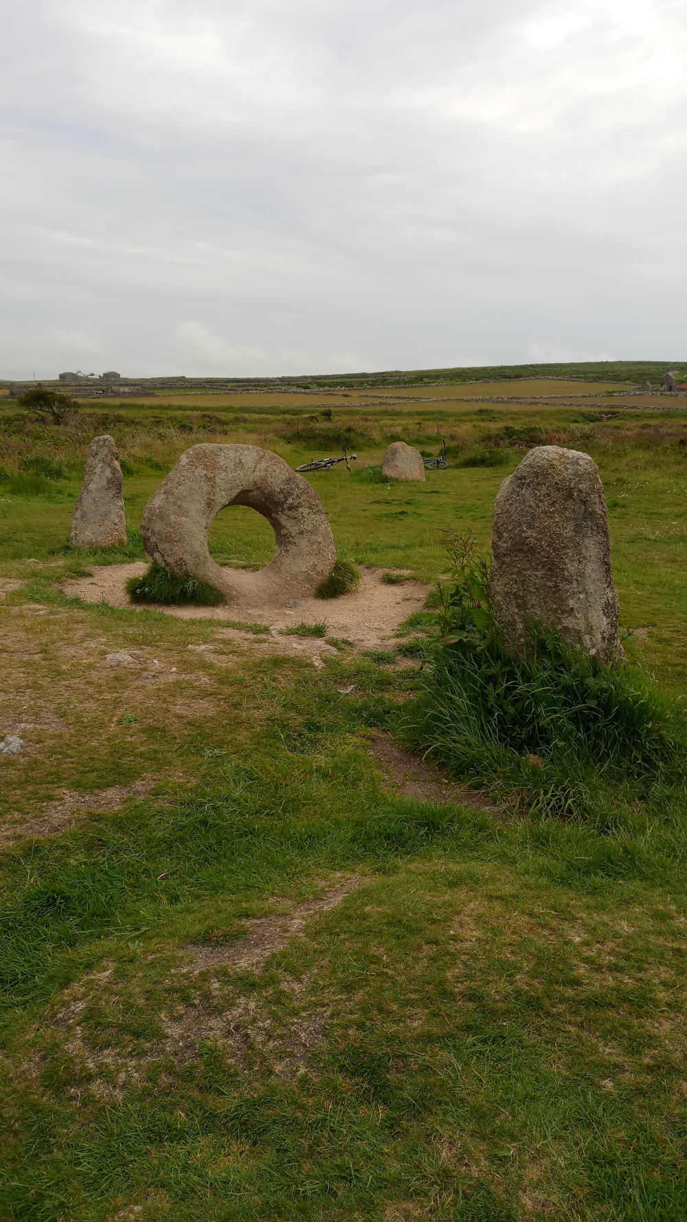 Stone circle, possibly a Neolithic or Bronze Age monument, set in a grassy field under a cloudy sky. The most prominent feature is a large, horizontally-placed stone with a roughly circular hole in its center. Other standing stones of varying heights are also visible within the circle. Two bicycles are casually leant against some of the stones in the background, suggesting a contemporary setting despite the ancient nature of the stones. The overall impression is one of peaceful ancient history juxtaposed against modern leisure.