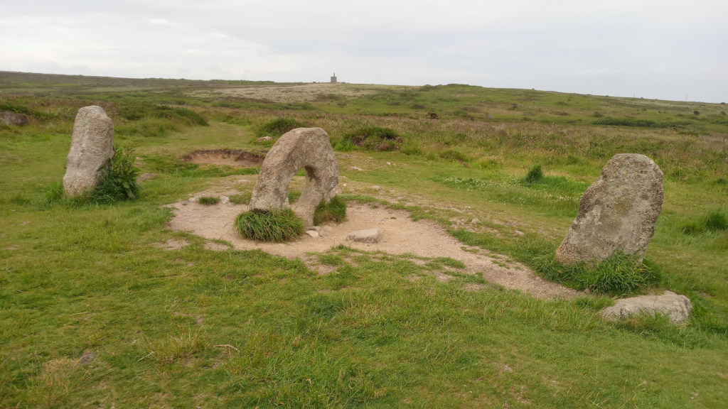 Landscape with several standing stones, possibly part of a Neolithic or Bronze Age monument.  One stone is shaped like a ring. The stones are set in short, grassy terrain, and a distant structure (possibly a tower or building) is visible on the horizon. The overall impression is one of ancient history and a somewhat desolate, yet peaceful, setting.