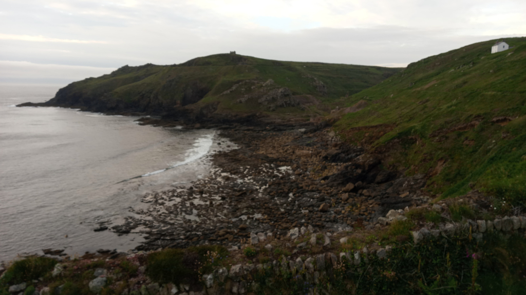 Coastal scene, likely in a region with a temperate climate. The foreground is composed of a rocky shoreline with low-lying vegetation and a partially visible stone wall. The mid-ground features a rocky beach extending into calm, grey-green water, with a small wave breaking near the shore. The background displays a verdant, hilly coastline leading up to a higher point with what may be some ruins or an old structure. A small, white building is visible on the upper right of the hill. The overall mood is serene and somewhat muted, due to the overcast sky.