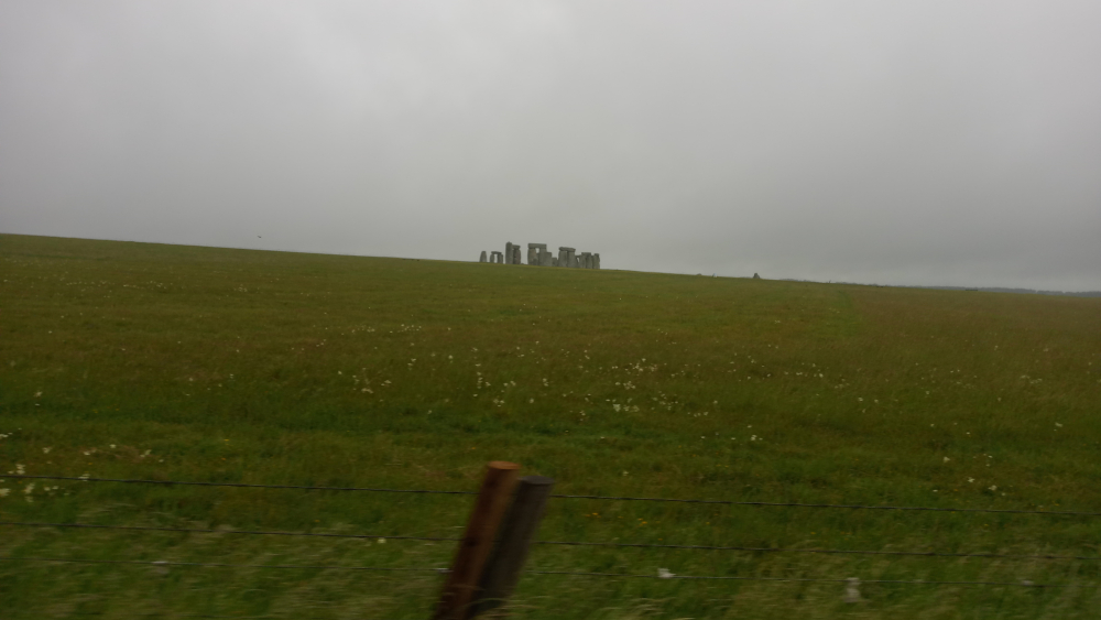Stonehenge in the distance, on a cloudy day. The foreground is a field of grass with a wire fence. The overall mood is muted and somewhat sombre due to the overcast sky and the distant view of the monument.