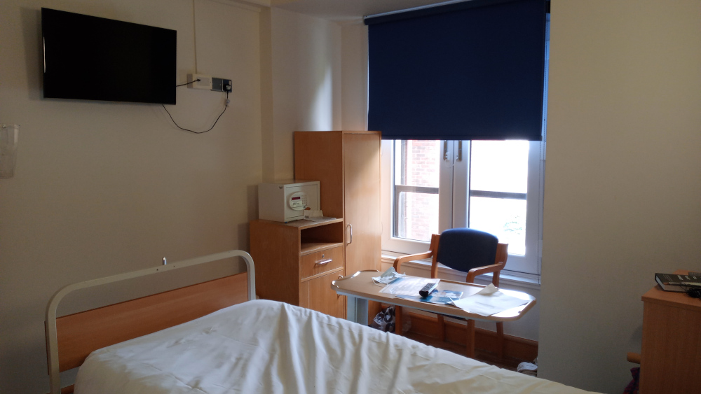 Sparsely furnished hospital room. The room contains a bed with white linens, a small television mounted on the wall, a wooden wardrobe, a small bedside table with a safe, a chair, a small writing desk with papers and a remote, and another small wooden cabinet. The overall impression is one of clean, functional simplicity. The window is covered by a dark blue blind, and sunlight is visible through the blind and window panes.