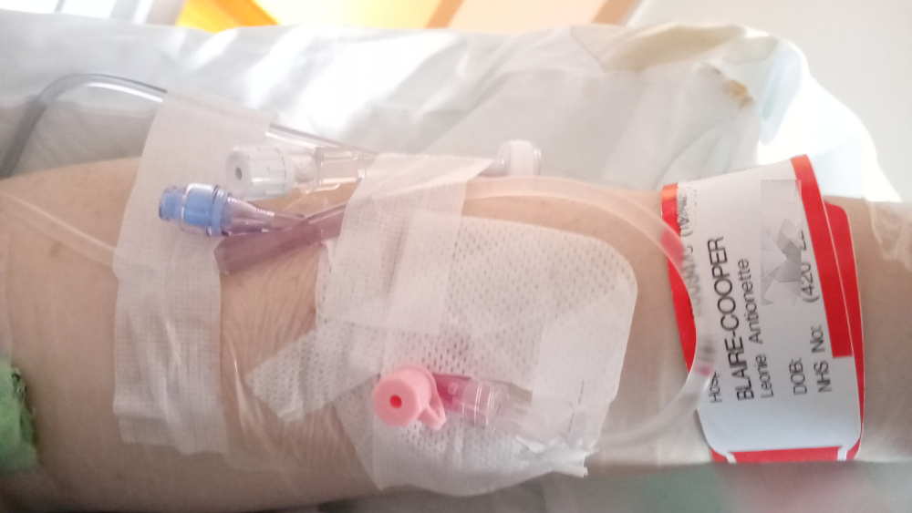 Close-up of Leonie's arm in a hospital bed, with an intravenous (IV) line inserted. Her arm is bandaged, and a hospital identification bracelet is visible. The overall impression is one of medical treatment and possible illness or injury.