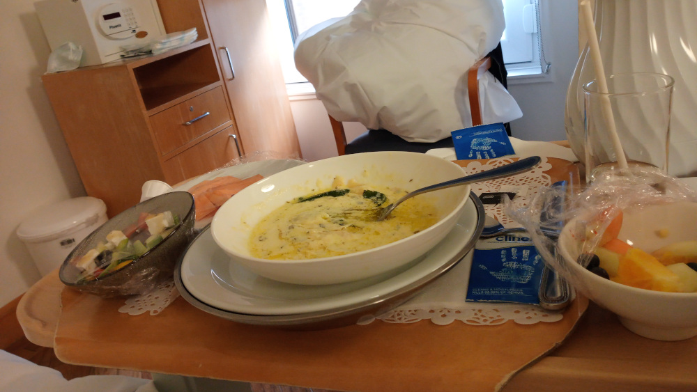 Hospital meal tray containing a partially eaten bowl of creamy soup, a small bowl of salad, a small bowl of fruit, and a piece of salmon. The tray is on a wooden table next to a bedside cabinet. The overall impression is one of a hospital room and a patient's simple yet somewhat unappetising meal. The partially consumed food suggests that the patient may not have a strong appetite.