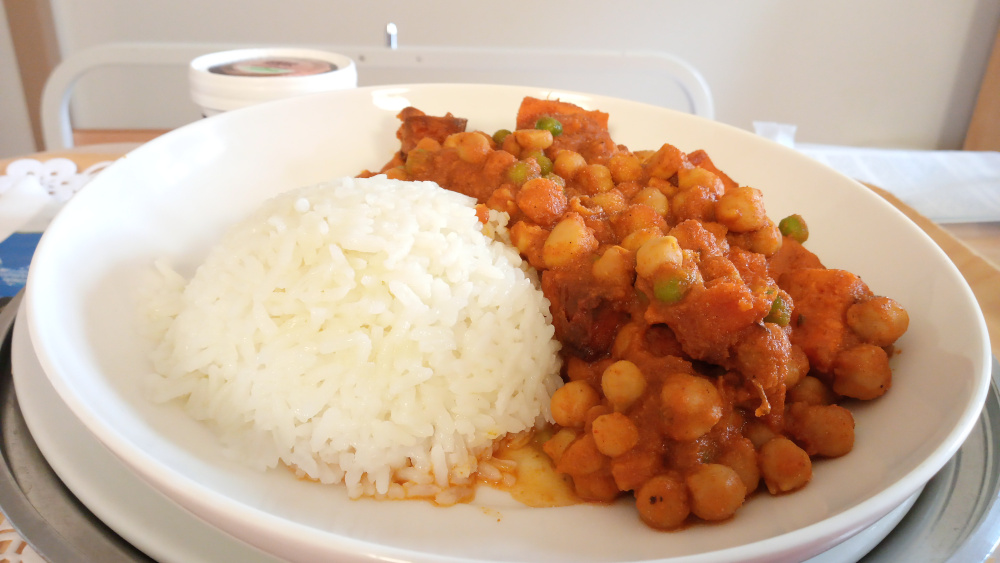 Plate of food containing a portion of white rice and a chickpea curry. The curry appears to include carrots or sweet potatoes and peas, and is a reddish-orange color indicative of a tomato-based sauce. The food is served on a white plate, which rests on a larger, possibly metallic, tray. A small container, possibly yogurt or a similar condiment, is visible in the background. The setting appears to be a hospital or medical facility, judging by the background elements.