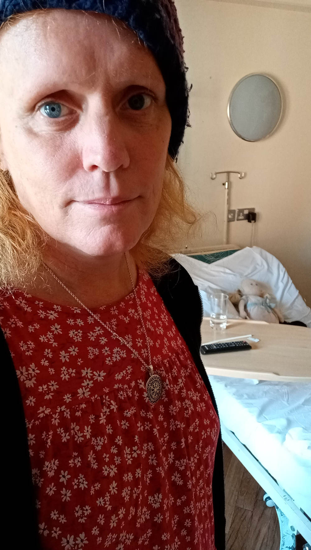 Leonie with shoulder-length reddish-blonde hair wearing a dark blue knitted hat and a red floral dress. She's also wearing a silver necklace with a circular pendant and is standing in what appears to be a hospital room. Part of a hospital bed and bedside table are visible in the background; a glass of water and a small stuffed animal are on the table. Her expression is serious and slightly sombre. The lighting and overall setting suggest a potentially contemplative or reflective mood, possibly related to a hospital stay or illness.