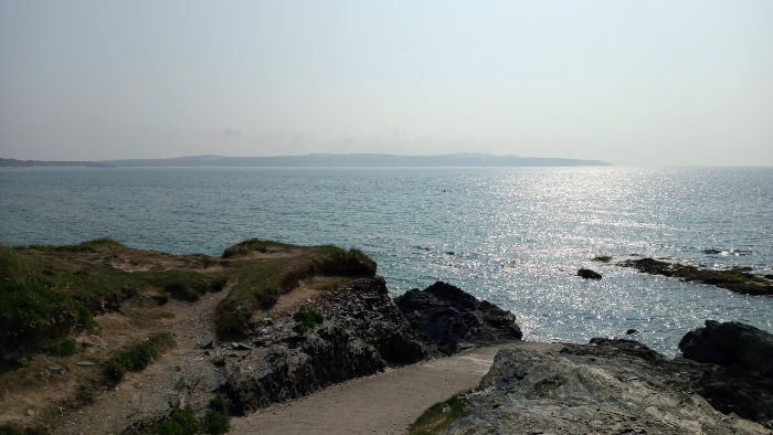 Tranquil coastal scene. From a slightly elevated vantage point, a rocky shoreline descends towards a calm sea sparkling in the sunlight. In the distance, a hazy landmass is visible on the horizon. The overall mood is peaceful and serene.