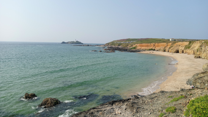 Tranquil coastal scene on a sunny day.  A calm, teal-coloured sea laps gently at a sandy beach, which curves gently around a rocky headland. In the distance, a small island with a lighthouse is visible. The cliffs are a mix of greyish-brown rock and reddish-brown earth, showing signs of erosion. The overall impression is one of serenity and natural beauty. A few people are visible on the beach, giving a sense of scale.