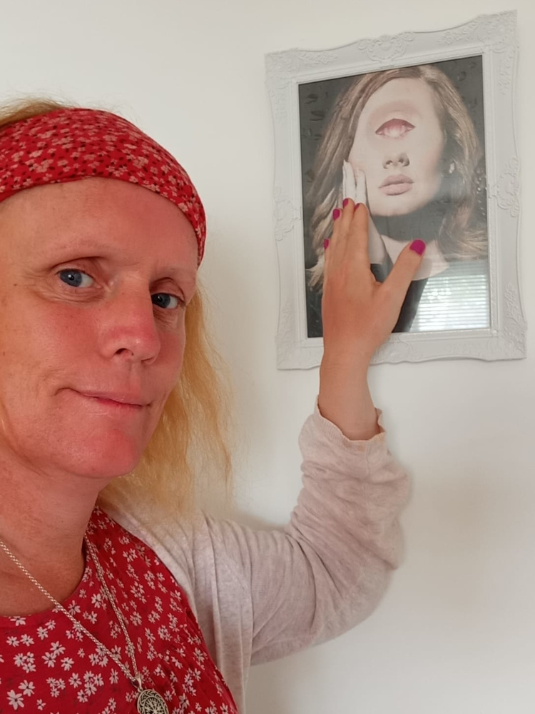 Leonie with blonde hair wearing a red floral headband and a red floral top, reaching out to touch a framed picture on a white wall. The picture depicts a woman with an eye replacing her forehead, creating a surreal and slightly unsettling effect. The woman in the photograph appears serene despite the unusual feature. The overall scene suggests a personal reflection or artistic appreciation of the surreal artwork.