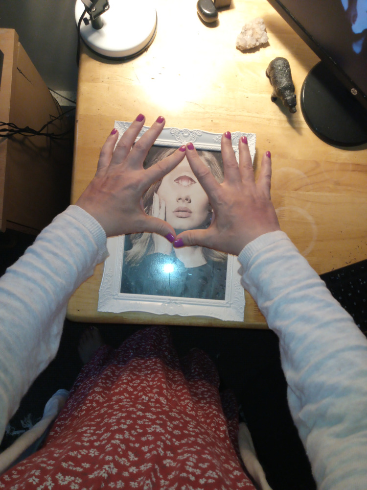 Leonie's hands forming a triangle shape around a framed picture of a woman. The woman in the picture appears to have a third eye in the centre of her forehead. The overall composition gives a mystical or surreal feel. The person's clothing and the items visible on the desk in the background suggest a setting related to spirituality or esoteric practices.