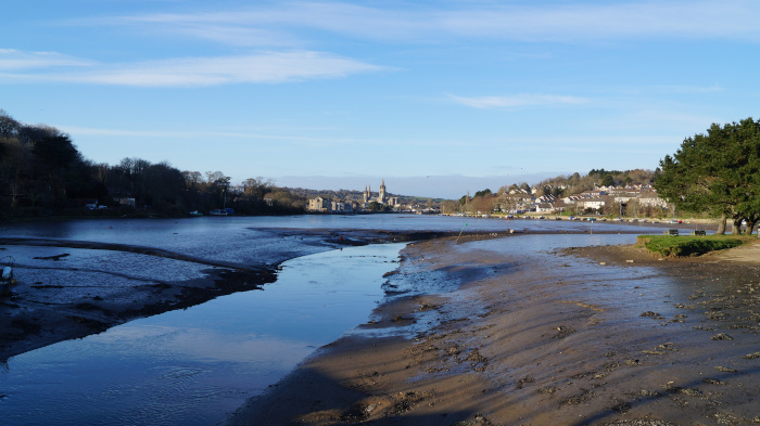 Fredelig landskap ved vannkanten, sannsynligvis ved lavvann. En rolig vannvei strekker seg ut og avslører en bred flate med mudderbanker i forgrunnen. På den andre siden av vannet er en by eller landsby synlig, med bygninger omgitt av trær, og en fremtredende kirke eller katedral er synlig midt i silhuetten. Himmelen er klar, lyseblå, noe som antyder et dagslys med godt vær.