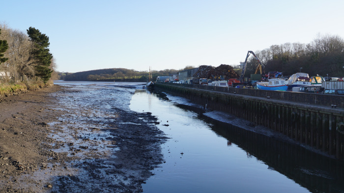 Utsikt over en vannvei ved lavvann. Forgrunnen domineres av synlige mudderbanker. På den andre siden av vannveien er en kai synlig, med båter fortøyd langs den og en betydelig haug med skrapmetall eller tømmer. Bak kaien er det trær og bygninger, noe som indikerer en noe industriell eller muligens maritim setting. Himmelen er klar og blå, noe som antyder et dagsscene.