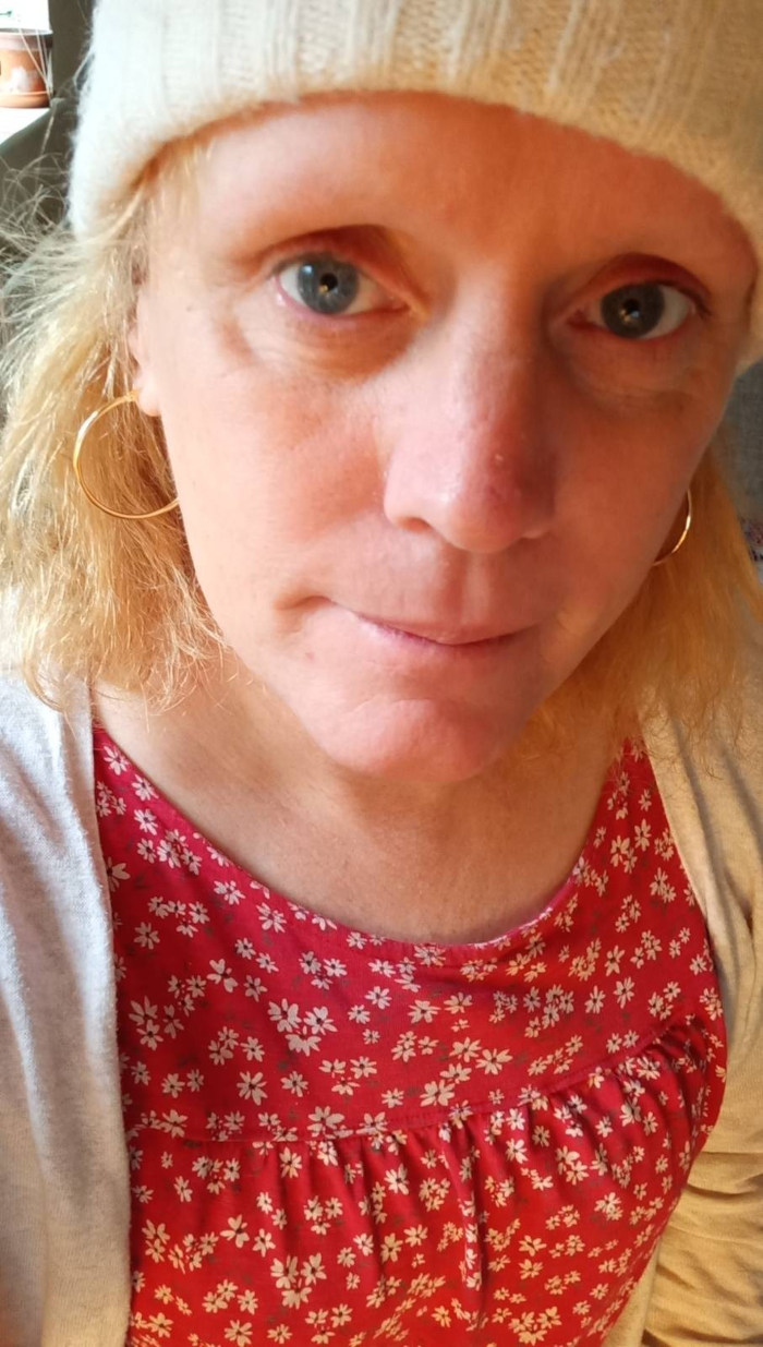 Nærbilde av en lyshudet kvinne med skulderlangt blondt hår. Hun har på seg en kremfarget strikket lue, gullfargede øredobber og en rød kjole med et lite hvitt blomstertrykk. Uttrykket hennes er nøytralt, nesten tankefullt. Belysningen antyder et innendørs miljø.