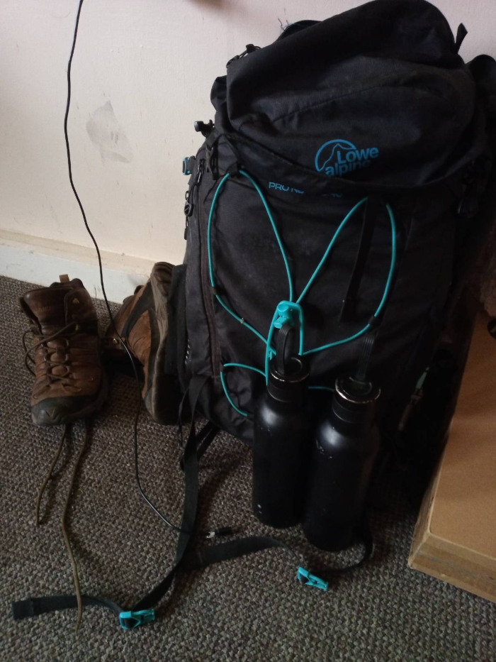 Svart Lowe Alpine-ryggsekk, to svarte vannflasker festet til sekken med blågrønne klips, og et par brune tursko som ligger på et teppebelagt gulv inntil en vegg. En svart kabel er også synlig og slynger seg over gulvet. Helhetsinntrykket er av utstyr forberedt for en fottur.