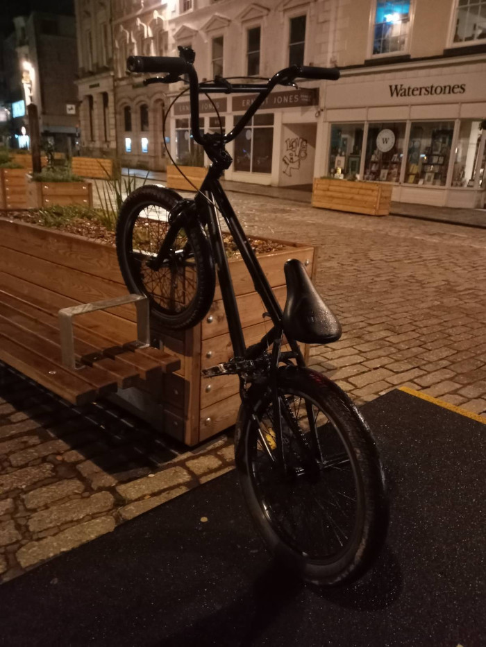 Svart BMX-sykkel lent mot en trebenk om natten. Sykkelen står parkert på et asfaltert område utenfor det som ser ut til å være en rekke butikker, hvorav en er en bokhandel i Waterstones. Scenen er svakt opplyst, noe som gir bildet en atmosfærisk, nattlig kvalitet.