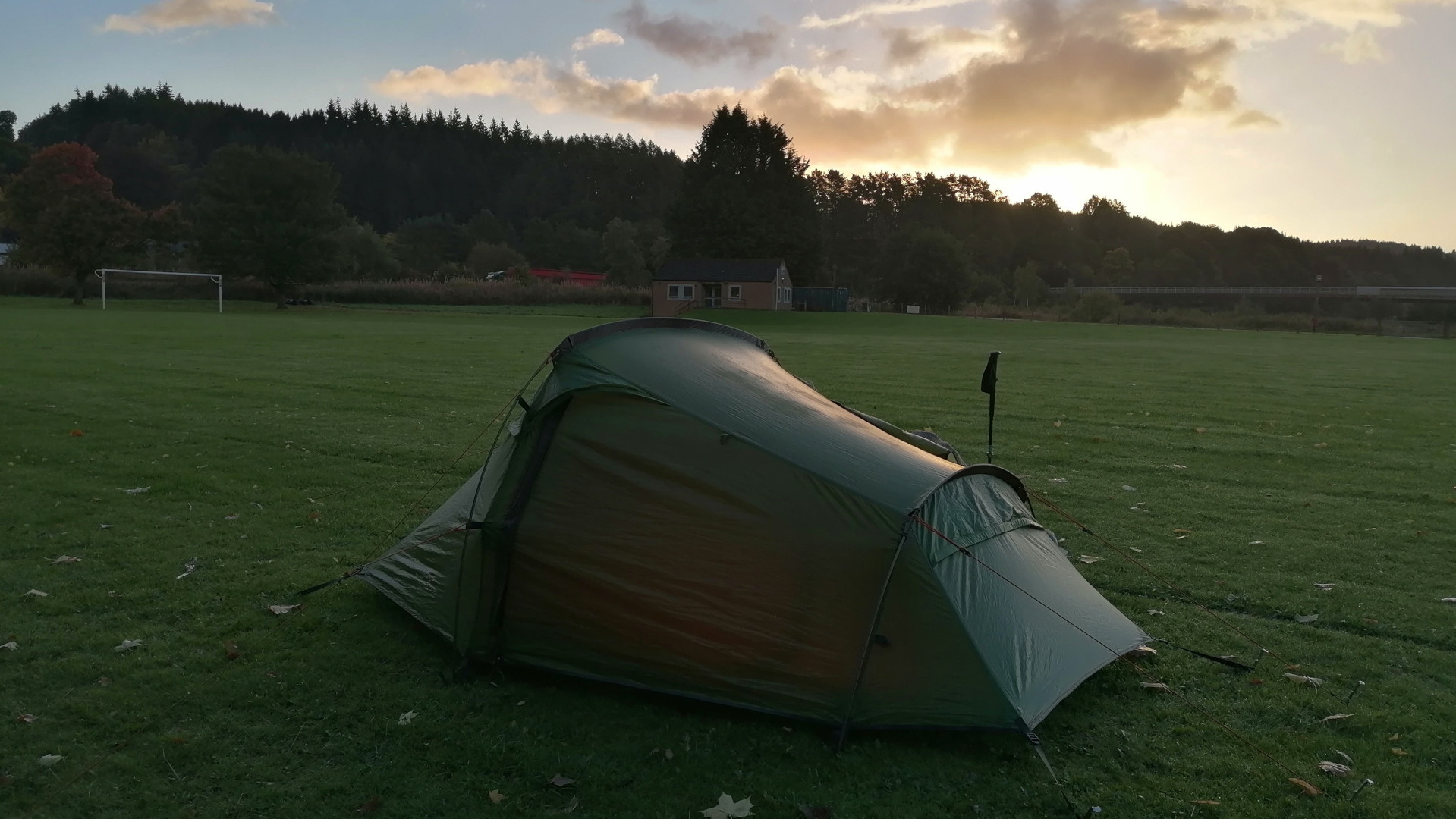 a dark-green tent pitched on a grassy field at sunset. The field appears to be near a wooded area and a small building is visible in the background. The sky is a mix of clouds and clear areas, with the sun setting behind the trees. The overall scene suggests a peaceful camping within an outdoor recreation setting.