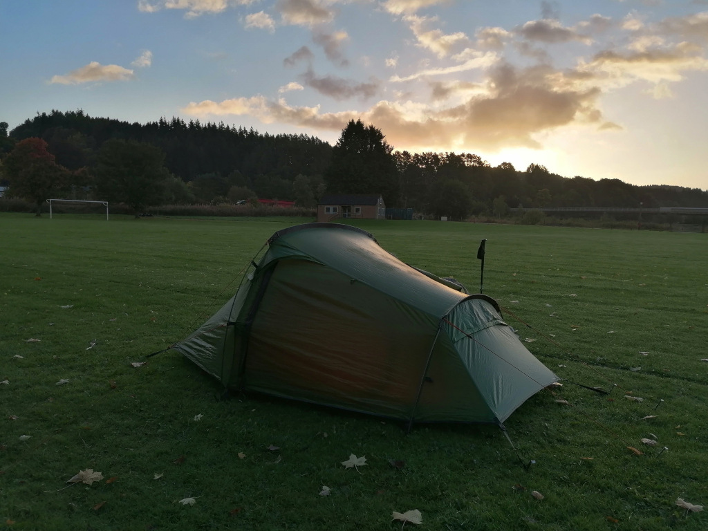 Dark-green tent pitched on a grassy field at sunset. The field appears to be near a wooded area and a small building is visible in the background. The sky is a mix of clouds and clear areas, with the sun setting behind the trees. The overall scene suggests a peaceful camping within an outdoor recreation setting.