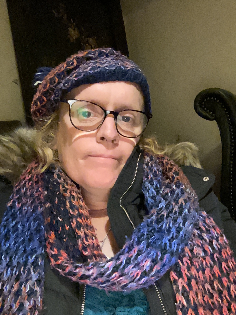 Leonie positioned slightly off-center, looking directly at the camera. Her expression is neutral, almost pensive. She appears to be sitting. Her hair is light blonde/brown, partially visible beneath the hat. Her glasses are dark-rimmed and appear to have a slightly reflective quality. The scarf is loosely draped around her neck, with the ends hanging down. Her jacket has a fur-like trim around the hood.