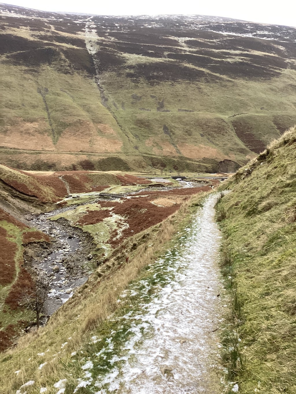 Snow-dusted trail winding along a hillside, overlooking a valley with a stream. The valley is characterised by its varied terrain, with patches of green and brown grass, and a small stream meandering through it. In the distance, a larger hill or mountain rises, showing layers of colour and texture. The scene evokes a sense of tranquil, desolate beauty typical of a sparsely populated countryside.