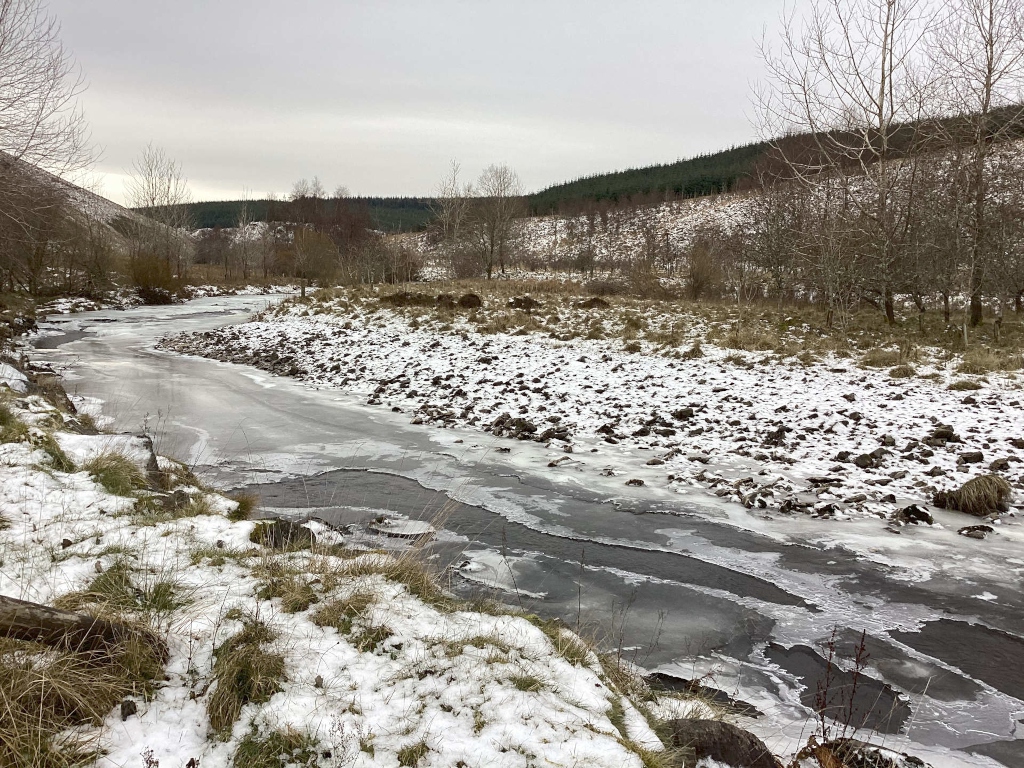 Partially frozen river meandering through a snow-dusted landscape. The riverbanks are rocky and sparsely vegetated, with leafless trees and bushes lining the banks and hills in the background. A light snow covers the ground, and the sky is overcast and grey. The overall mood is serene yet cold.