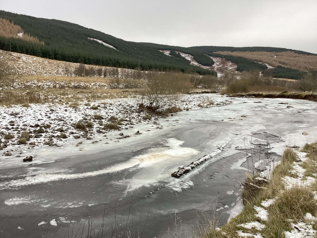 Partially frozen river flowing through a wintry landscape. The river is dark, with patches of ice and snow along its banks and surface. The banks are sparsely vegetated, with some dry grasses and shrubs visible. In the background, there are dark green hills covered with trees, hinting at a rural or mountainous setting. A log is partially submerged and frozen into the river.