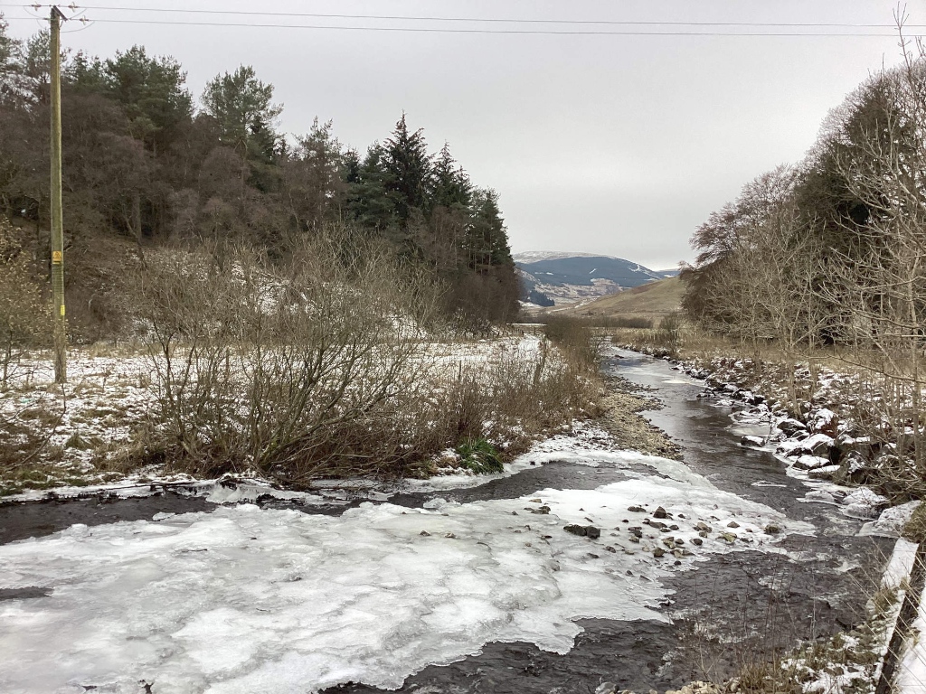 Partially frozen stream or river winding through a snowy landscape. The river is flanked by leafless trees and bushes, indicating a winter setting. In the background, there's a line of hills under a grey, overcast sky. A utility pole is visible on the left side of the frame. The overall impression is one of a peaceful, cold, and somewhat desolate winter scene.