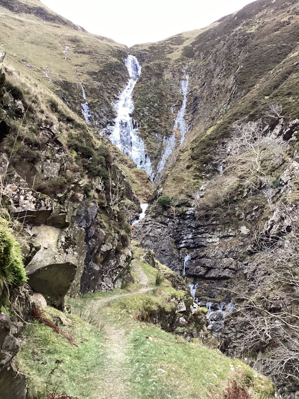 Partially frozen waterfall cascading down a rocky mountainside. A trail meanders along the base of the cliff face, leading towards the waterfall. The overall atmosphere is one of cold, rugged beauty, typical of a winter landscape. Sparse vegetation clings to the rocky slopes. A small figure, possibly a hiker, is visible near the base of the falls, providing a sense of scale.