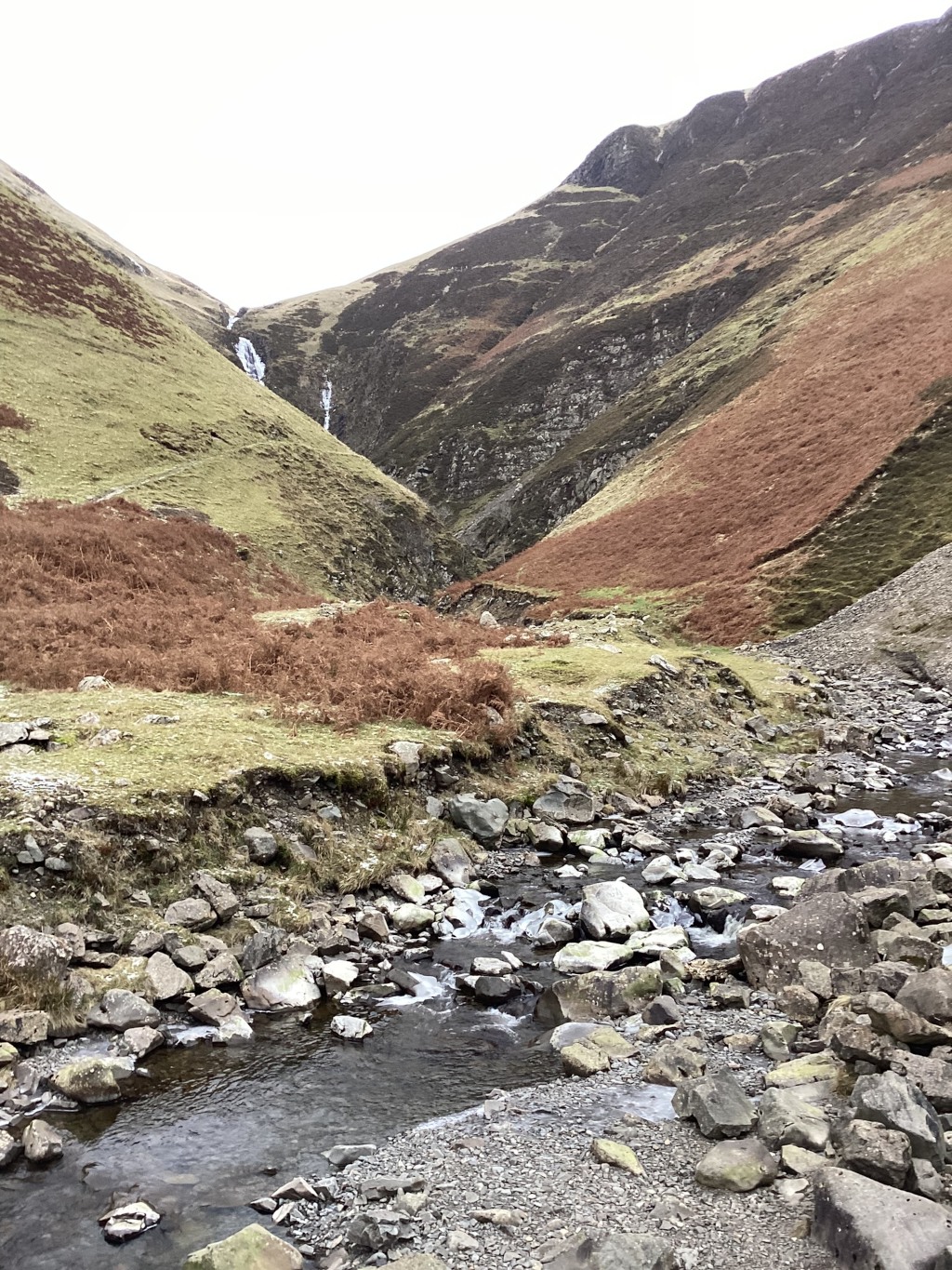 Rocky stream flowing through a valley between two mountains. The mountainsides are a mix of greens and browns, with some patches of reddish-brown vegetation visible. A waterfall is visible in the distance, cascading down the mountainside into the valley. The overall impression is one of a wild, rugged, and somewhat desolate, yet beautiful landscape.