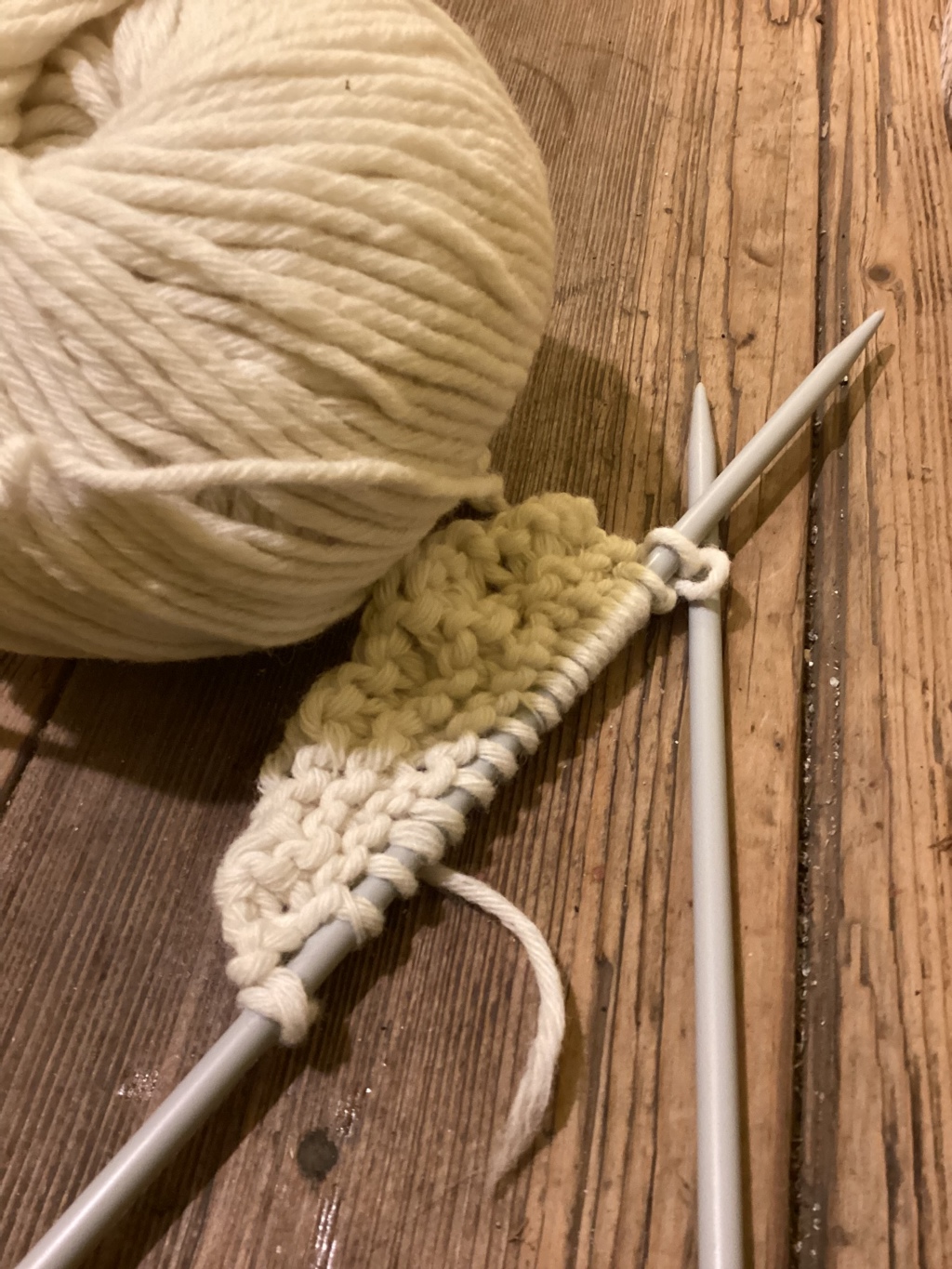 Ball of off-white yarn and knitting needles with a small piece of knitting in progress. The knitting shows a subtle colour change from a pale yellow/cream to the off-white of the main yarn ball. The setting appears to be a rustic wooden surface.