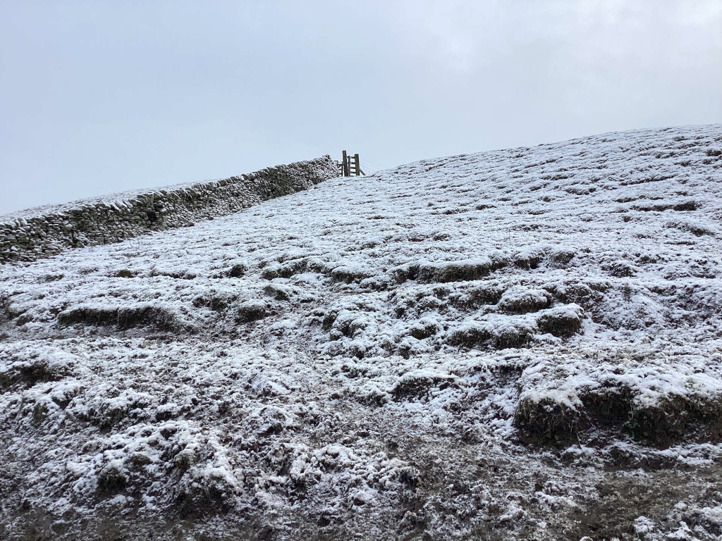 Snow-covered hillside with a low stone wall running along its lower edge. A simple wooden stile is visible in the wall. The scene is minimalistic, with a muted colour palette dominated by the whites and greys of the snow and stone. The overall impression is one of quiet, wintry solitude.