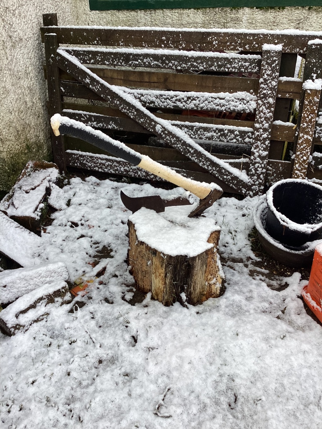 Large axe stuck in a tree stump. The scene is outdoors, covered in a light layer of snow. A wooden fence, also covered in snow, is visible in the background. There are some pieces of wood and a black pot on the ground nearby. The overall impression is one of a rustic, possibly rural setting, perhaps in the winter.