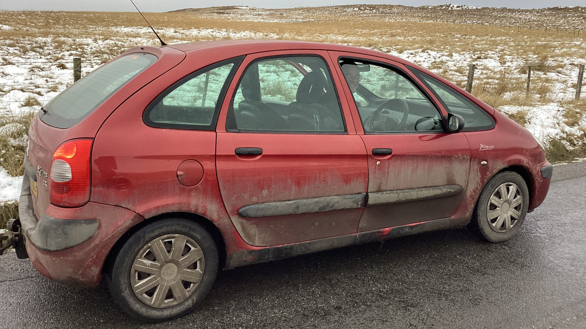 A dirty, red Citroën Xsara Picasso parked on the side of a road. The car is covered in mud and dust. The background is a bleak, wintry landscape of brown fields with patches of snow. The overall impression is one of a journey, perhaps a long one, in harsh conditions.