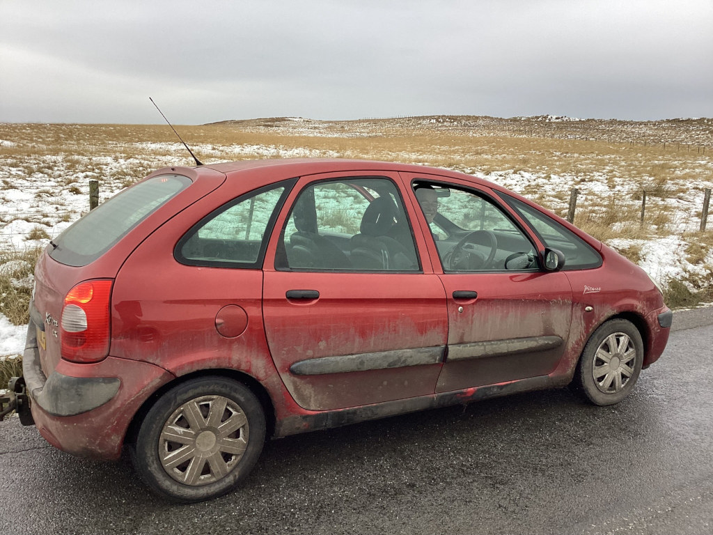 Dirty, red Citroën Xsara Picasso parked on the side of a road. The car is covered in mud and dust. The background is a bleak, wintry landscape of brown fields with patches of snow. The overall impression is one of a journey, perhaps a long one, in harsh conditions.