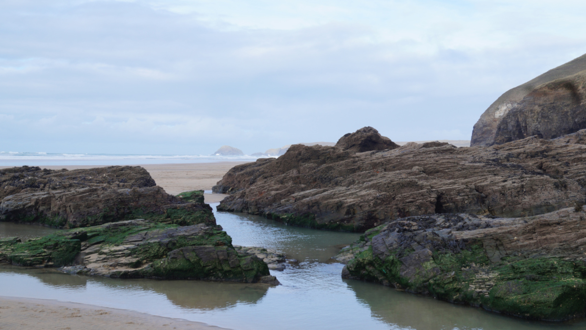 A tranquil coastal scene. Dark, layered rocks, partially covered in green algae, form natural pools and channels filled with calm, shallow water. A sandy beach and a calm ocean are visible in the distance under a cloudy sky. The overall impression is one of serene, natural beauty.