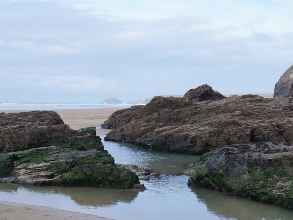 Tranquil coastal scene. Dark, layered rocks, partially covered in green algae, form natural pools and channels filled with calm, shallow water. A sandy beach and a calm ocean are visible in the distance under a cloudy sky. The overall impression is one of serene, natural beauty.