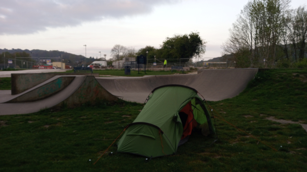 Green two-person tent pitched on the grassy area next to a skate park. The skate park features concrete ramps and a slightly overcast sky is visible in the background. A person in a bright yellow jacket is visible in the distance, seemingly near the skate park. The overall impression is of a somewhat desolate or quiet scene, perhaps suggesting camping or temporary shelter in an unconventional location.