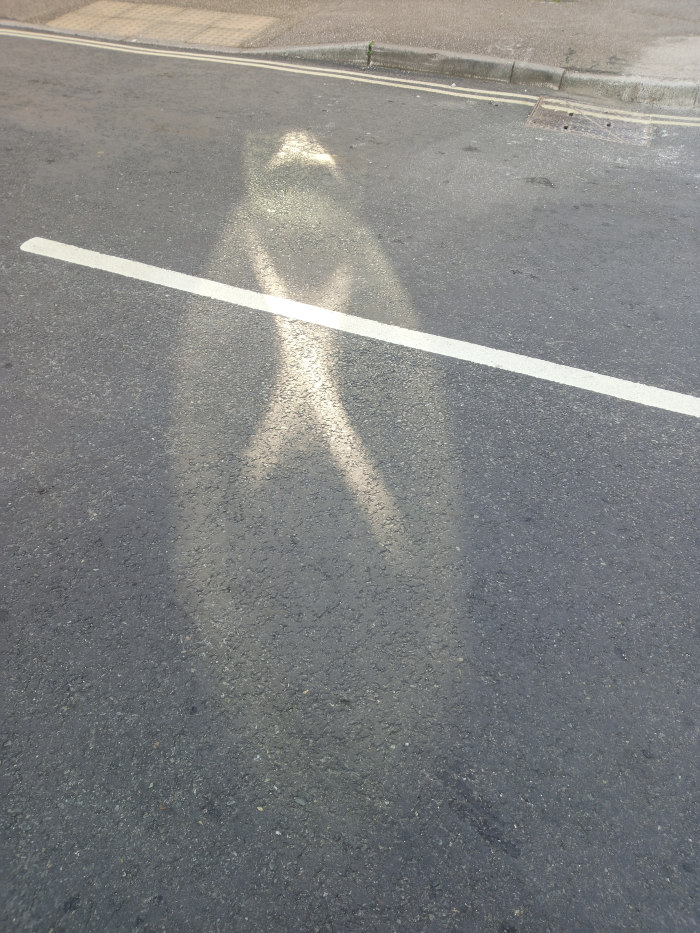 Section of asphalt road with a white line painted across it. A blurred, elongated shadow, suggestive of a person, is cast onto the road surface by sunlight. The shadow is distorted and somewhat ethereal due to the angle of the sun and surface reflectivity.