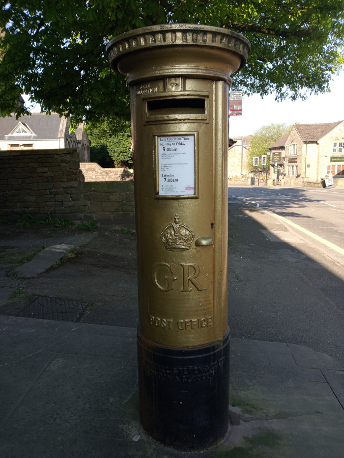 gold-painted post box on a street corner. The box is typical of a traditional British design, with the royal cypher GR visible. A small sign on the box displays collection times. The background includes a quiet street scene with buildings and trees.