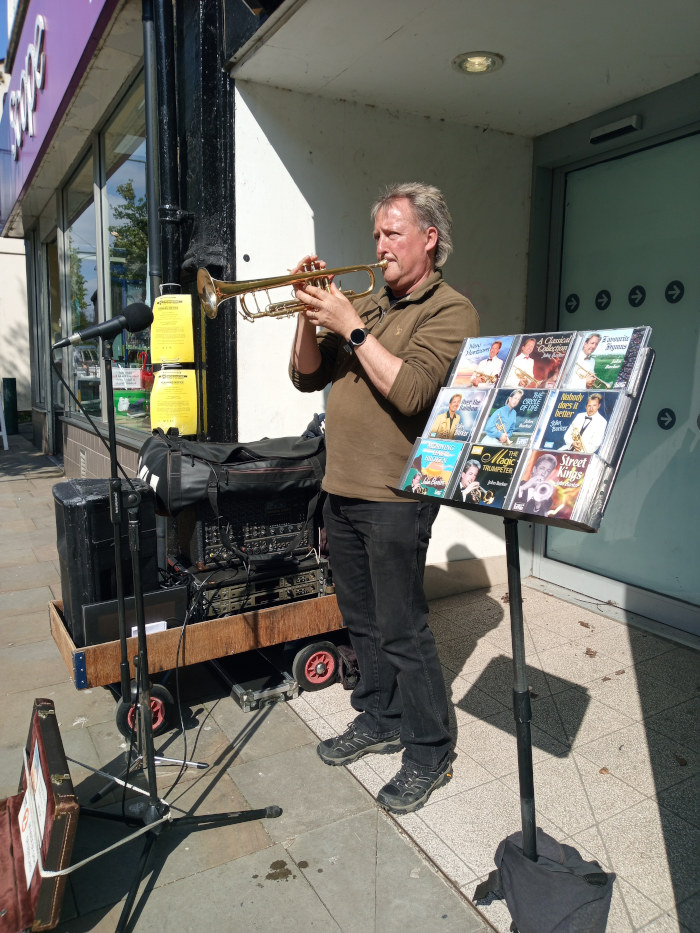 Middle-aged man busking on a sunny day. He's playing a trumpet, and his equipment—amplifier, microphone, and a small wooden cart—is neatly arranged beside him. A music stand holds a collection of CDs, suggesting he might be selling his music or promoting it.