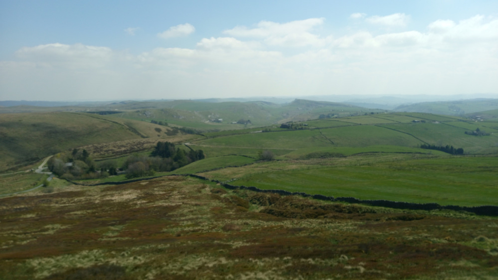 Rolling green hills and valleys, dotted with patches of darker vegetation and the occasional farm building. A winding road is visible in the mid-ground, leading towards a more densely wooded area. The overall mood is one of tranquil serenity and vastness. The sky is mostly clear and bright, contributing to the feeling of a calm, sunny day.