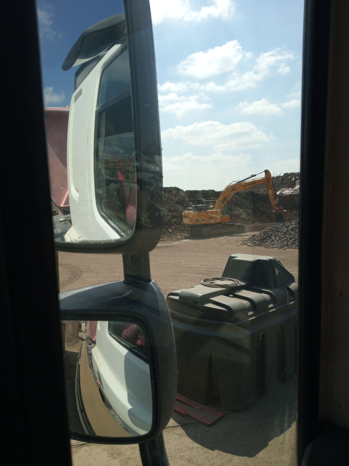 View from inside a truck cab, looking out through the driver's side mirrors. The mirrors reflect a construction site, with a large yellow excavator the focal point. A dark gray or black storage container is present in the foreground, also viewed through the truck's windows. A partially visible pink object is seen in both the mirrors.