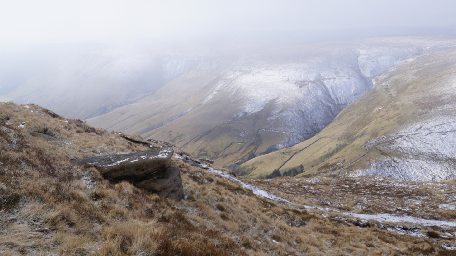 A panoramic view of a snow-dusted mountain landscape.  The foreground features a rocky outcrop covered in sparse, dry-looking vegetation. The middle and background consist of rolling hills and valleys, some patches of snow are visible on the hillsides. The overall atmosphere is cold, somewhat misty, and serene, conveying a sense of vastness and isolation.