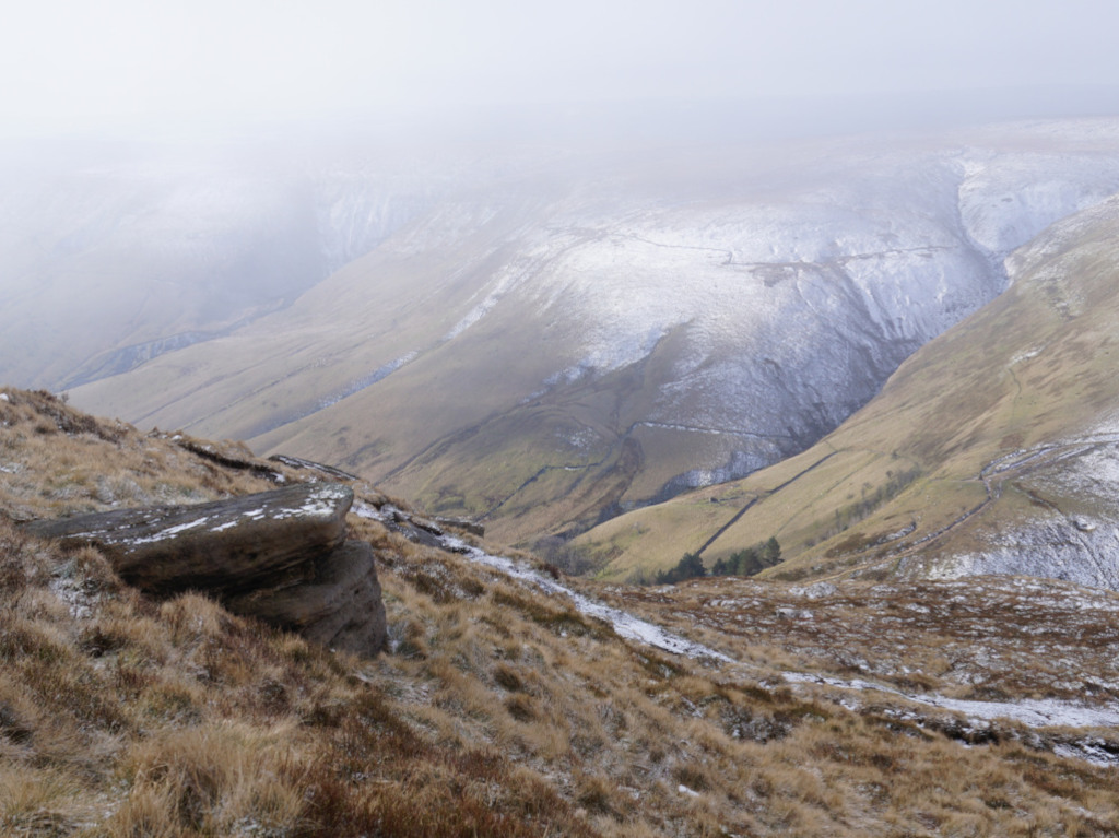 Panoramic view of a snow-dusted mountain landscape.  The foreground features a rocky outcrop covered in sparse, dry-looking vegetation. The middle and background consist of rolling hills and valleys, some patches of snow are visible on the hillsides. The overall atmosphere is cold, somewhat misty, and serene, conveying a sense of vastness and isolation.
