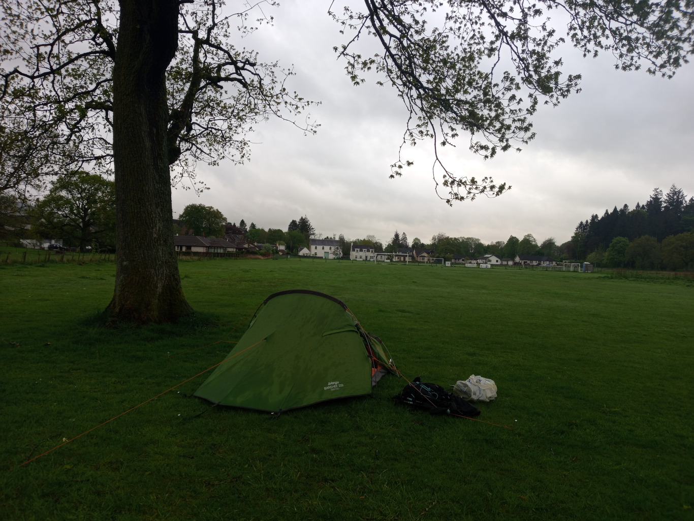 Olive-green dome tent, pitched on a lush, short-cropped green field. It's a slightly damp-looking tent, suggesting recent rain or dew, with subtle texture visible in the fabric. Beside the tent, partially obscuring the base, lies a dark-colored backpack and a lighter-colored, possibly grey or off-white, smaller bag or pack. A thin, orange-toned tent guy line stretches diagonally from a tent peg towards the left of the frame. No people are visible.