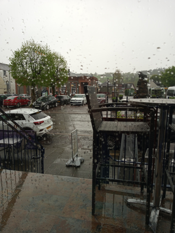 Inside a building, looking out through a rain-streaked window. The focus is on a wet, paved area outside with parked cars. The foreground is dominated by the out-of-focus image of patio furniture – a dark-colored wicker chair and metal table set. These appear wet from the rain, and the rain droplets distort their appearance. Part of a metal frame or stand also occupies the foreground. A small, grey A-frame sign stand is near the base of the window.  A staircase is partly visible to the left of the furniture.