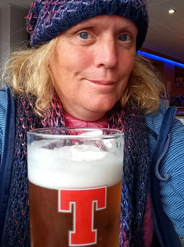 Enters on a middle-aged woman with shoulder-length, light blonde hair. She's wearing a dark blue and purple knitted beanie pulled low on her forehead, obscuring some of her hairline.  A chunky, multicolored knitted scarf, predominantly dark blue, purple, and hints of pink, is wrapped around her neck. She is dressed in a blue zip-up jacket.  Her expression is neutral, almost pensive, with a slight downward curve of her lips, creating a mildly melancholic yet calm mood. The woman is holding a pint glass of beer that appears to be a light-colored ale with a thick, creamy white head. The glass prominently displays a large red T logo, indicating a specific brand. She's slightly turned toward the viewer, creating a close-up intimate perspective.